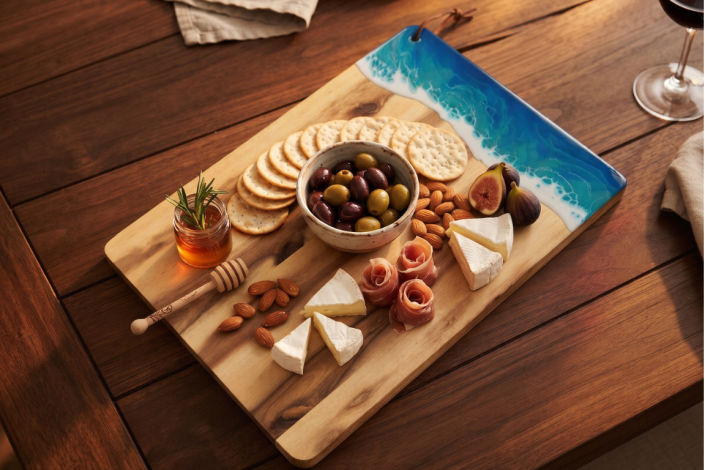 Servierbrett aus Akazienholz mit Meereseffekt aus Epoxidharz auf Holztisch. Auf dem Servierbrett sind verschiedene Snacks, wie Käse, Schinken, Feigen, Nüsse, Kräcker und ein Schälchen mit Oliven. Auf dem Tisch ist ein Rotweinglas und Stoffservietten in be