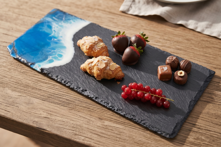 Schieferplatte mit Meereseffekt aus Epoxidharz auf einem Holztisch. Auf der Schieferplatte befinden sich süße Leckereien, wie Johannisbeeren, Pralinen usw.. Seitlich im Hintergrund ist ein beigefarbenes Tuch angedeutet