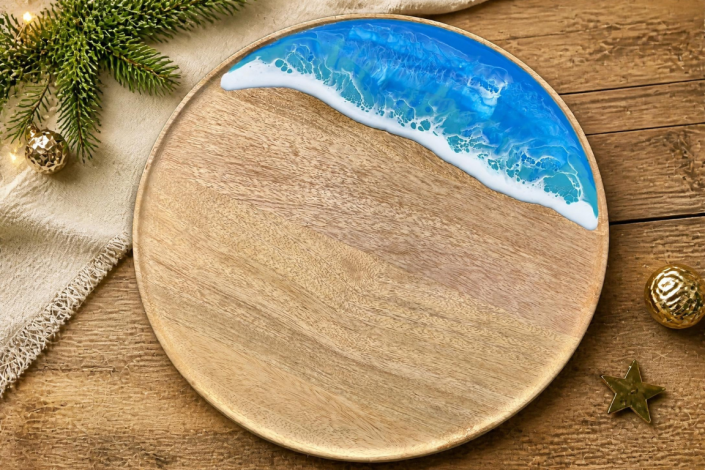 rundes Tablett aus Mangoholz mit Meereswellen-Effekt aus Epoxidharz, auf einem Holztisch, auf dem teilweise eine beigefarbene Tischdecke liegt, sowie etwas weihnachtliche Deko wie ein Tannenzweig mit Lichterkette, goldene Weihnachtskugeln und ein Stern