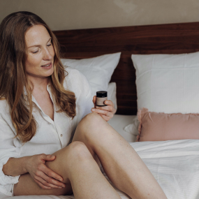 Eine Frau sitzt auf dem Bett und hält ein Glas Rosas Deocreme in der Hand
