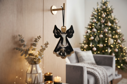Engel aus Epoxidharz in schwarz und gold aufgehängt mit einem schwarzen Satinband an einem goldfarbenen Haken an einer Holzwand. Der Wohnraum ist dekoriert mit einem grauen Sessel, modernen Kerzen in schwarz und weiß, einer Vase mit Eukalyptuszweigen und 