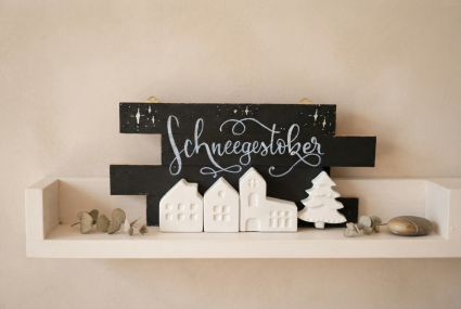 Eine Produktpräsentation des handgefertigten Deko-Objekts „Schneegestöber“. Das schwarze Holzschild steht auf einem hellen, cremefarbenen Holzregal vor einer warmen, beigen Wand. Auf den verschetzten Holzleisten des Schildes sind unten drei kleine, weiße 