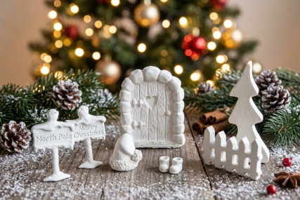 Ein handgemachtes, weißes Wichteltür-Set aus Keramik vor einem unscharfen Weihnachtsbaum mit Lichtern. Das Set enthält eine verzierte Tür, einen Zaun, eine Tanne, Wichtelstiefel, eine Mütze und zwei Hinweisschilder mit den Aufschriften „North Pole“ und „M