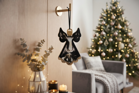 Engel aus Epoxidharz in schwarz und gold aufgehängt mit einem schwarzen Satinband an einem goldfarbenen Haken an einer Holzwand. Der Wohnraum ist dekoriert mit einem grauen Sessel, modernen Kerzen in schwarz und weiß, einer Vase mit Eukalyptuszweigen und