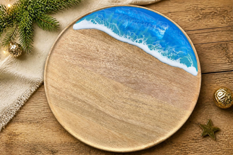 rundes Tablett aus Mangoholz mit Meereswellen-Effekt aus Epoxidharz, auf einem Holztisch, auf dem teilweise eine beigefarbene Tischdecke liegt, sowie etwas weihnachtliche Deko wie ein Tannenzweig mit Lichterkette, goldene Weihnachtskugeln und ein Stern