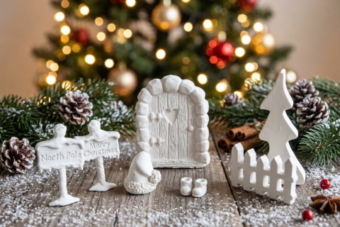 Ein handgemachtes, weißes Wichteltür-Set aus Keramik vor einem unscharfen Weihnachtsbaum mit Lichtern. Das Set enthält eine verzierte Tür, einen Zaun, eine Tanne, Wichtelstiefel, eine Mütze und zwei Hinweisschilder mit den Aufschriften „North Pole“ und „M