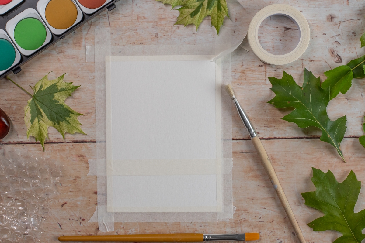 Ein Blatt Papier ist auf eine Unterlage geklebt - Pinsel, Tuschkasten und Blätter liegen daneben.
