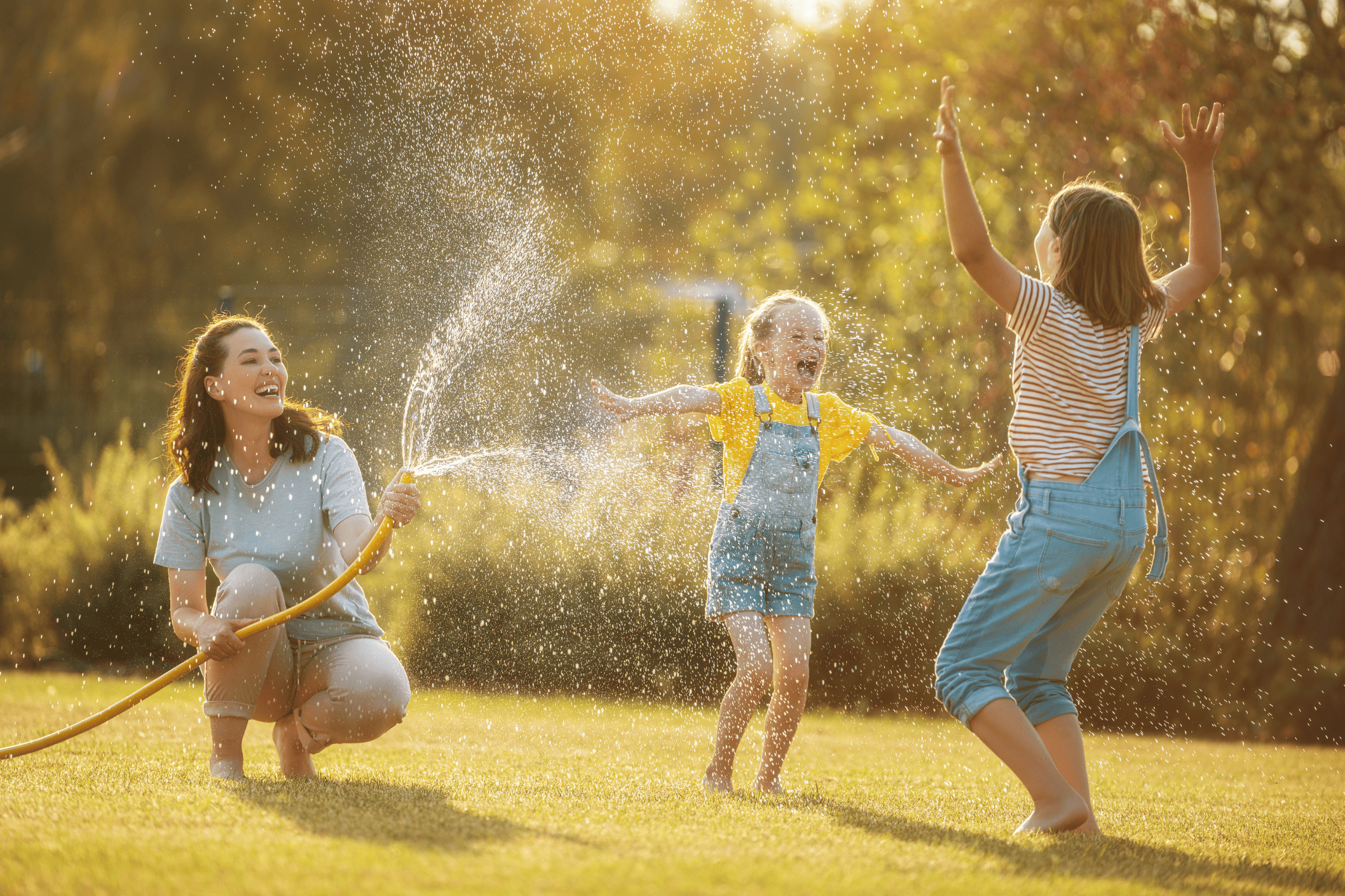 Eine Mutter spritzt ihre beiden Kinder mit dem Wasserschlauch im Garten nass.